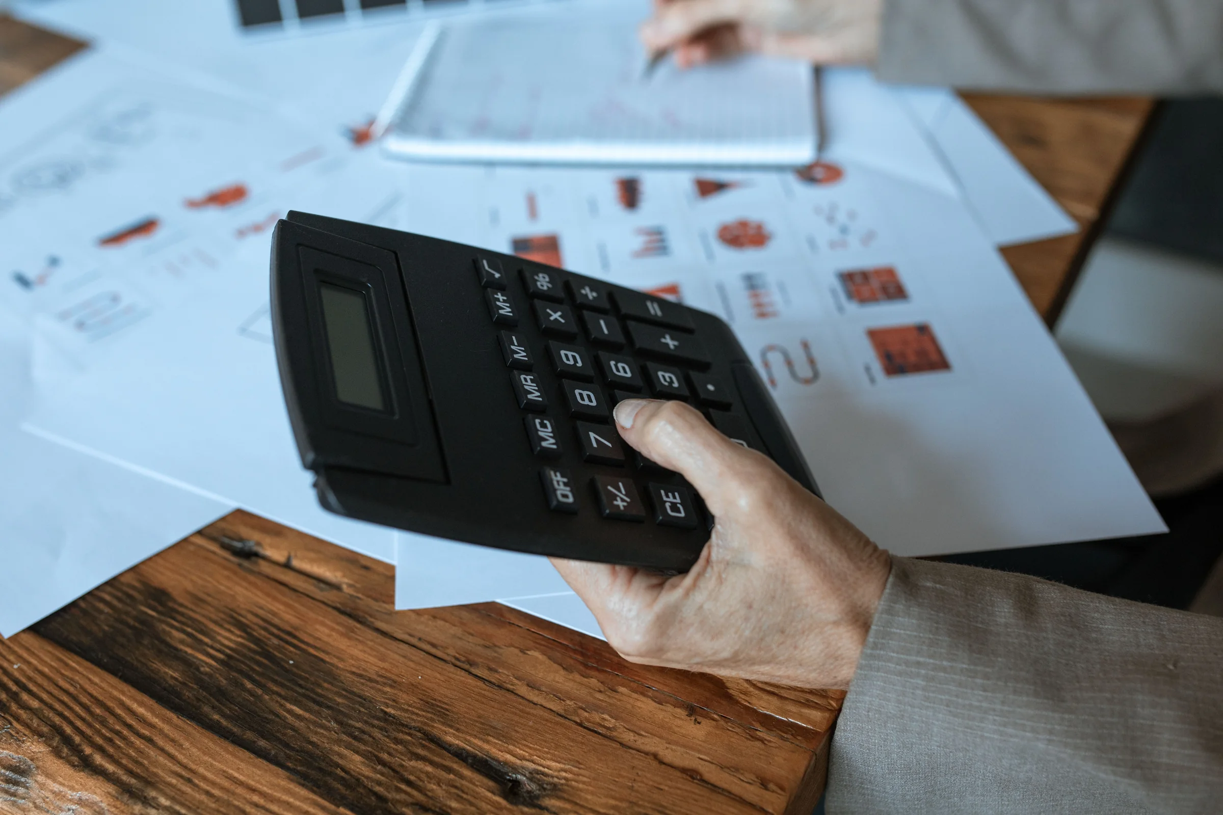 Mão utilizando calculadora diante de planilhas e gráficos financeiros.